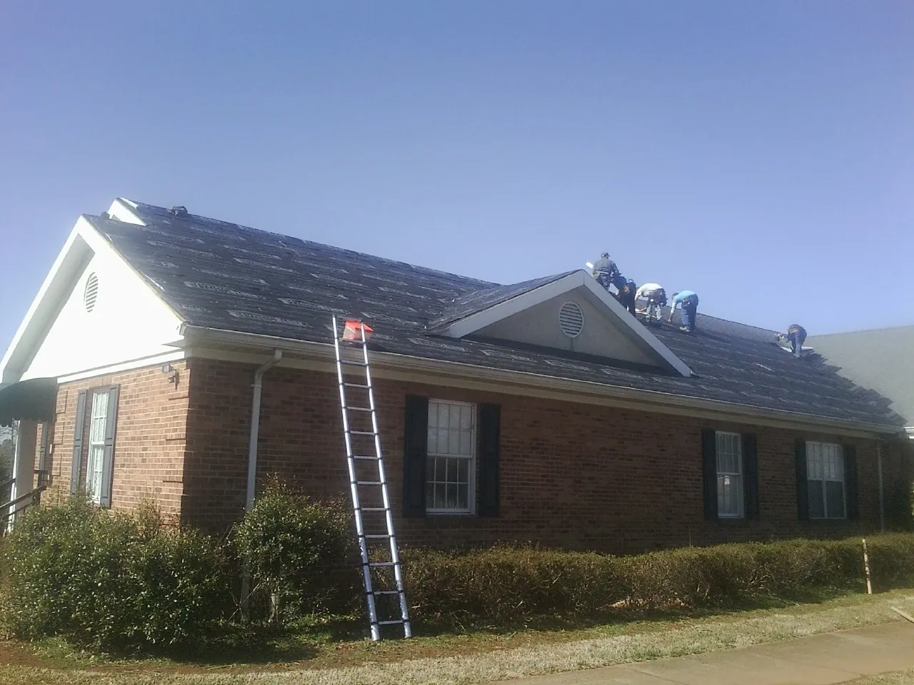 Crew tearing shingles off a brick ranch roof with a ladder set — Emergency Roof Repair work by our crew in Gilman